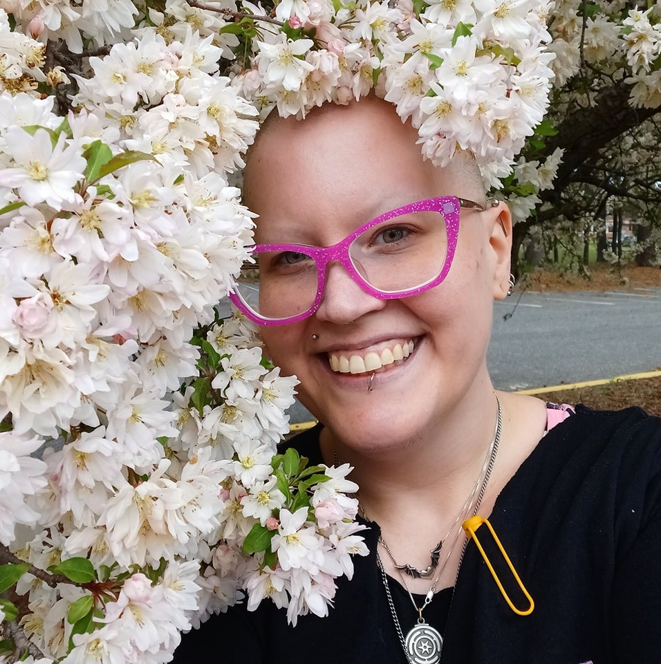 Lou, a nonbinary person with pink glasses and a shaved head, smiles broadly while posing under a tree in bloom with white flowers.